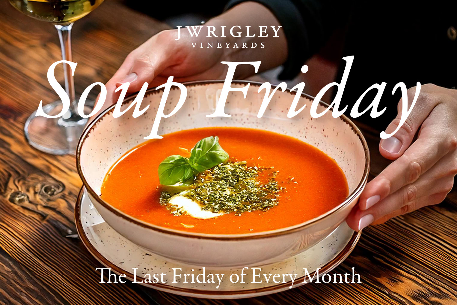 A bowl of tomato soup garnished with basil and herbs, with a glass of wine in the background, for Soup Friday at J Wrigley VY.